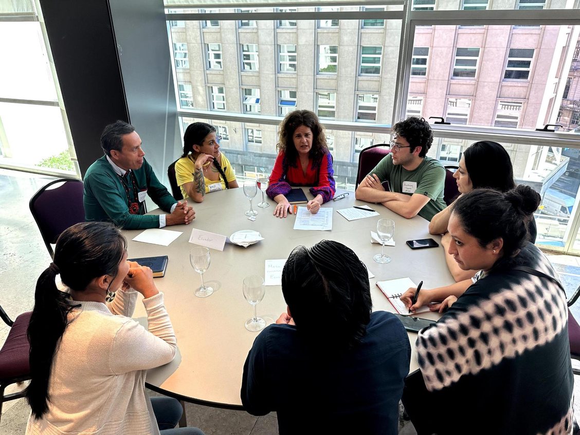 Anat Shenker-Osorio wearing a red top seated at a round table surrounded by organizers listening and taking notes.